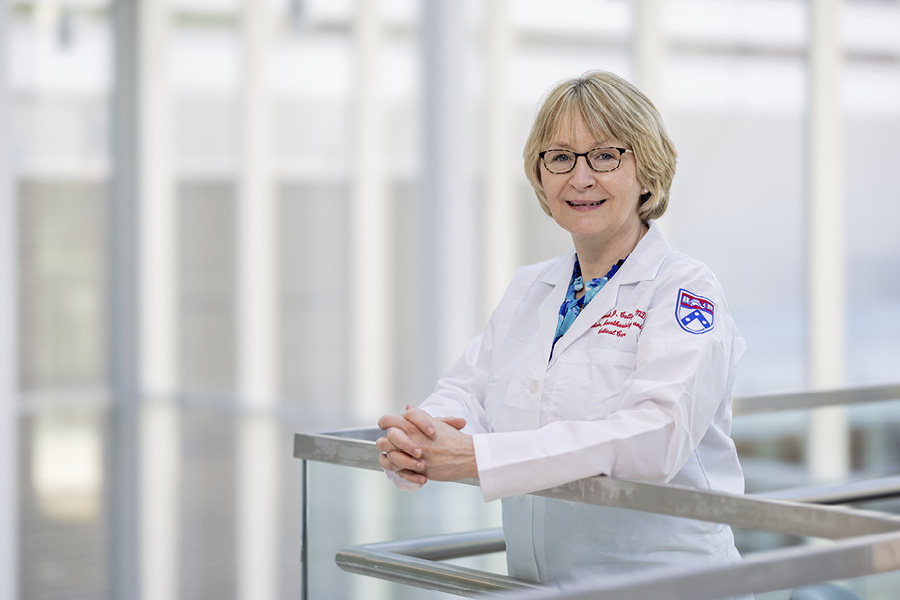 Deborah Culley, MD, chair of Anesthesiology and Critical Care, wears a white coat and smiles with her hands folded and resting atop a glass railing.